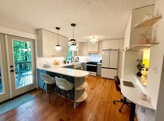 Bright modern kitchen with white island, pendant lights, wood floors, and glass doors opening to greenery