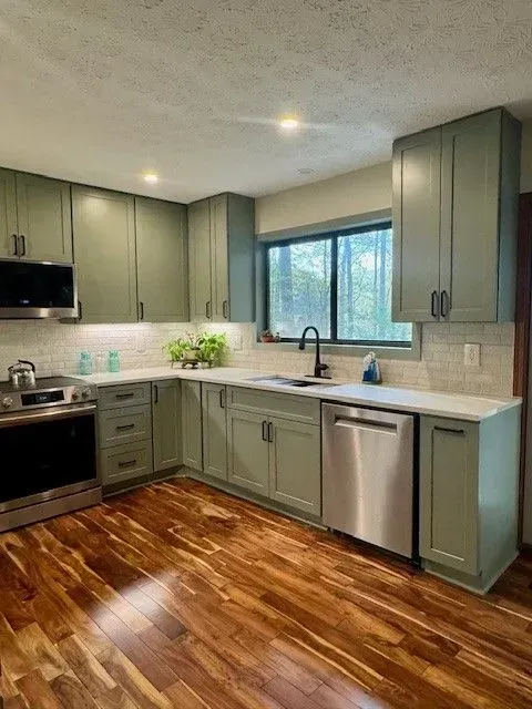 Modern kitchen with green cabinets, stainless appliances, wood floor, and large window over the sink