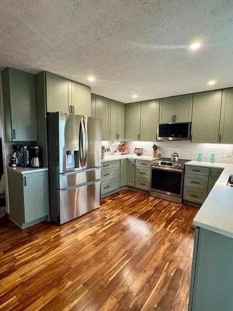 Modern kitchen with sage green cabinets, stainless steel appliances, and glossy hardwood floors.