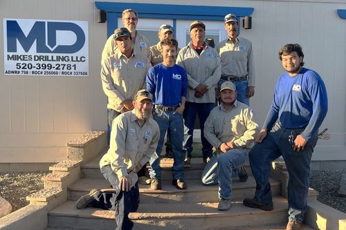 Group of workers in blue and tan shirts posing on outdoor steps beside a drilling company sign.