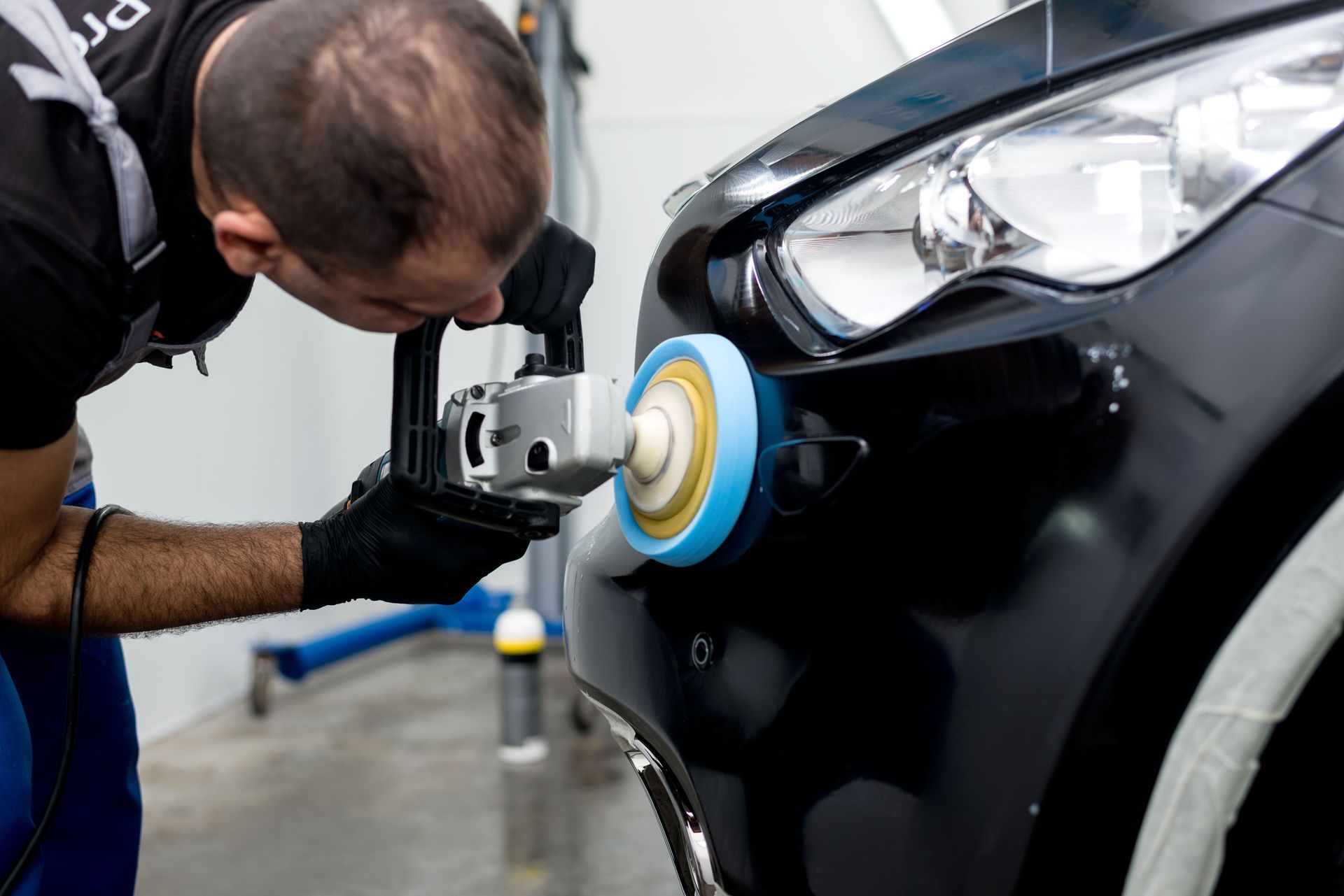 A person polishing a black car bumper with an electric buffer in a garage.