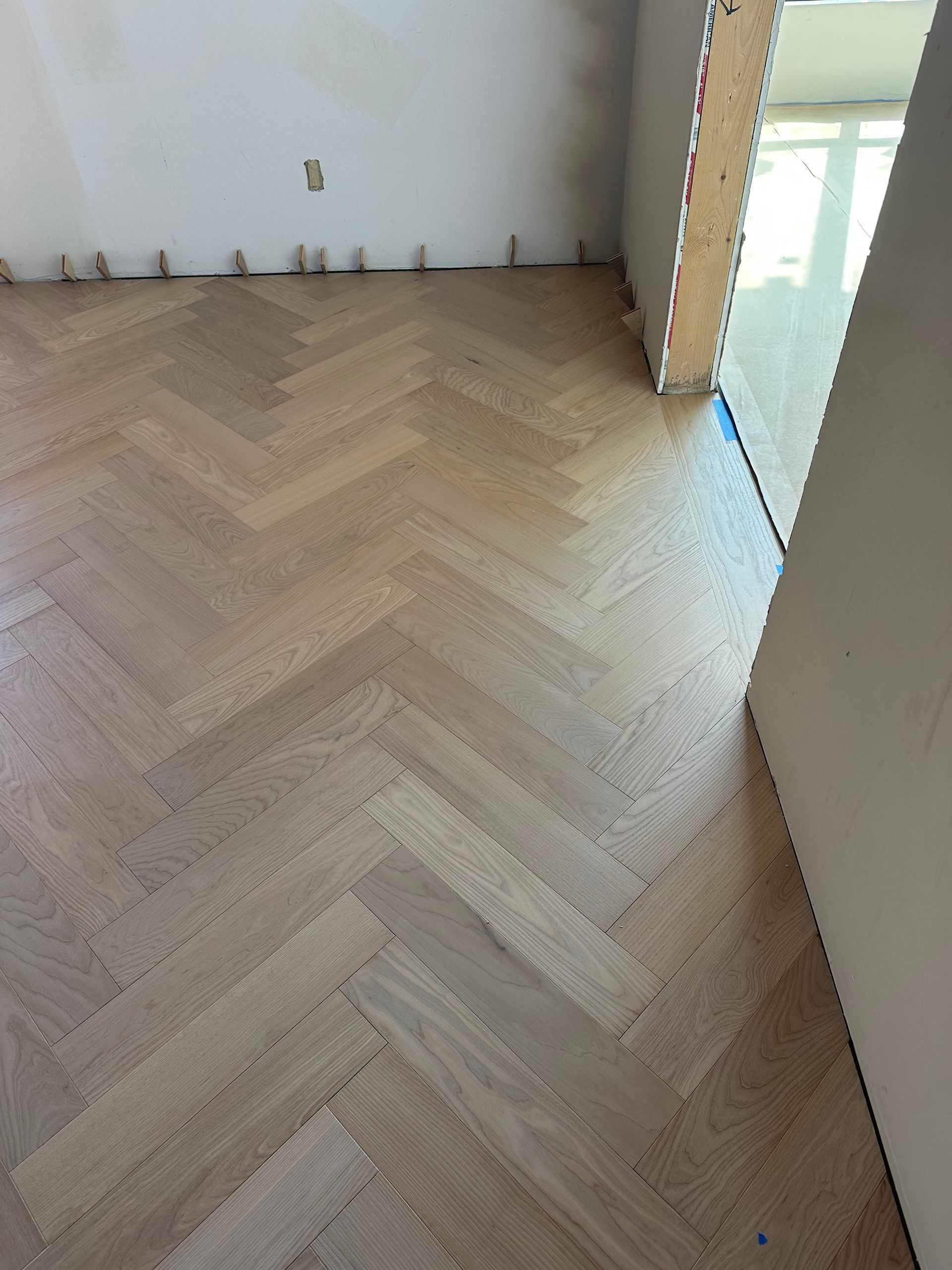 Herringbone pattern wood floor with natural light and visible unfinished wall.