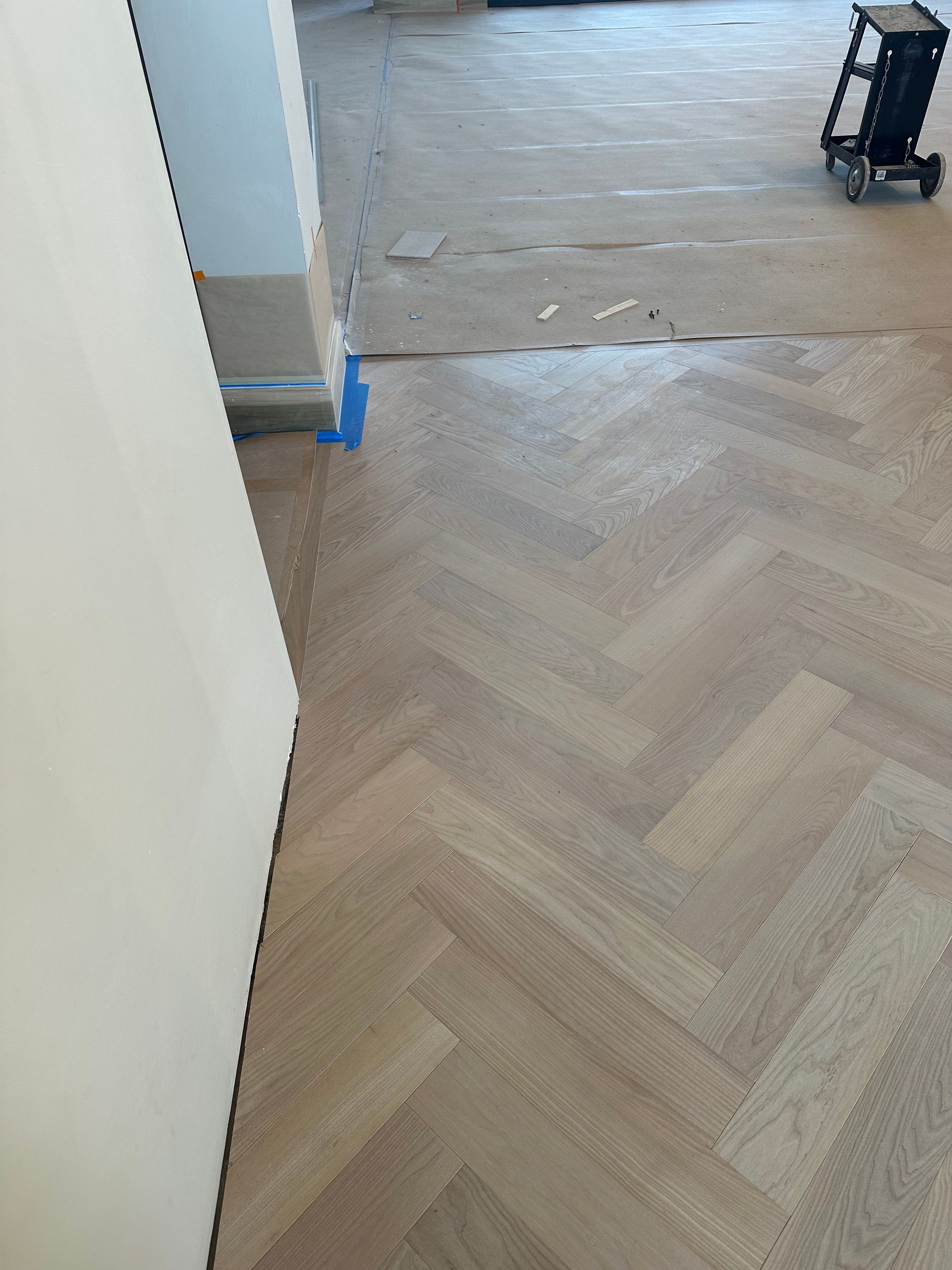 Herringbone wood floor, unfinished, next to a white wall.  Blue tape on wall near doorway.