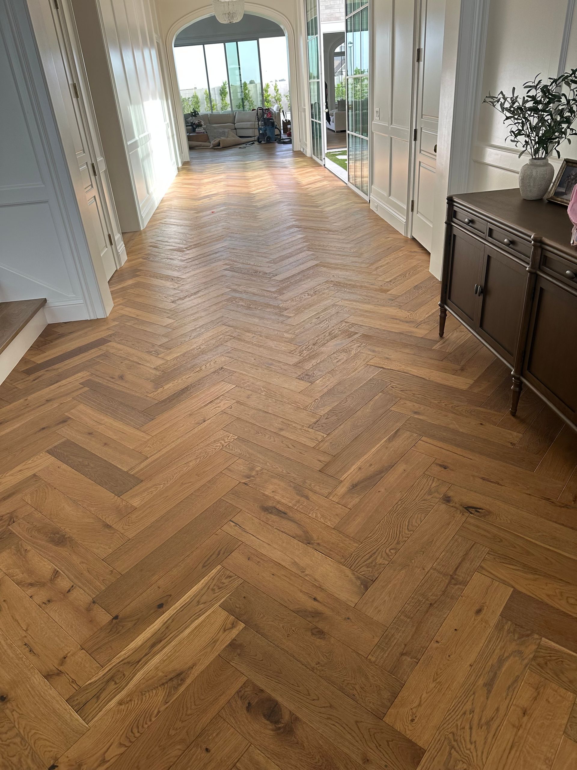Herringbone hardwood floor in a hallway, with a wooden cabinet on the right side and a light-filled doorway at the end.