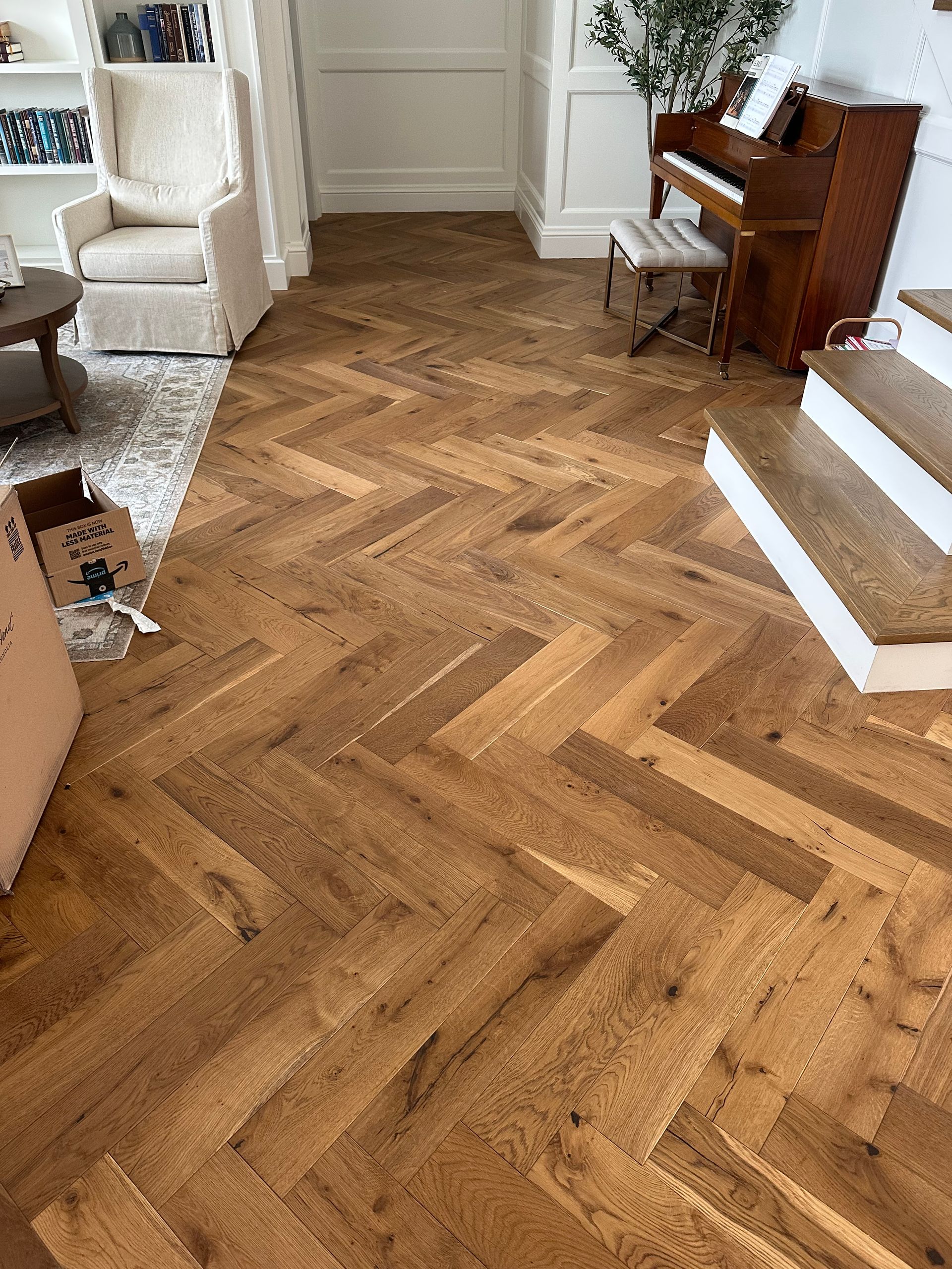 Herringbone-patterned hardwood floor in a living room with a piano, stairs, and a white armchair.