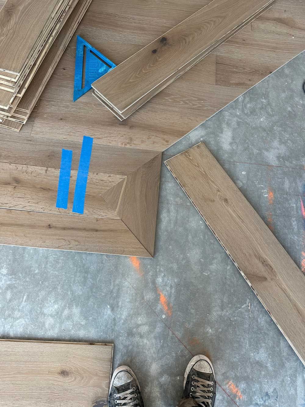 Person's feet near wooden planks on concrete floor with blue tape and a square.