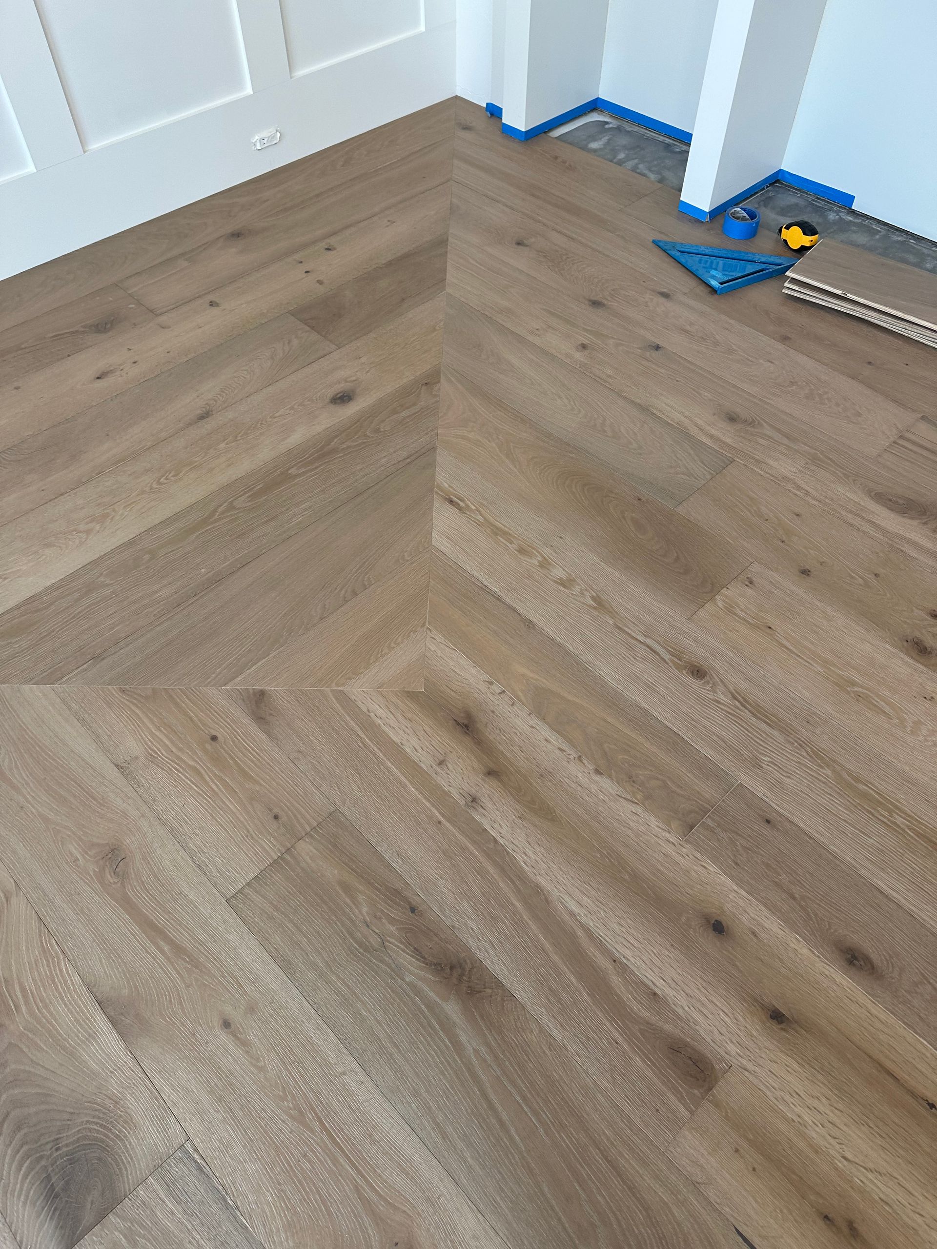 Herringbone patterned light wood floor in a room corner, with white trim and blue painter's tape.