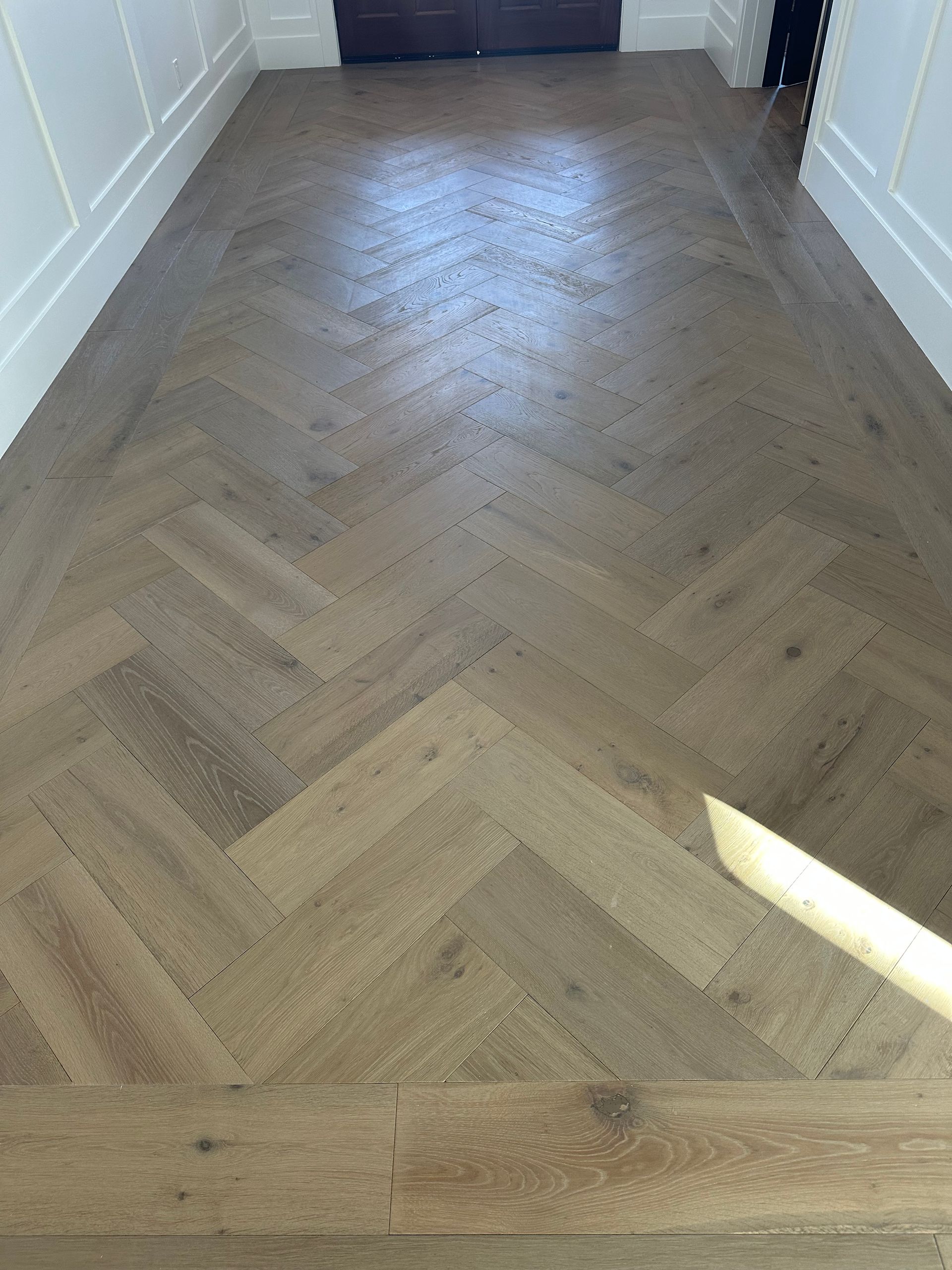 Herringbone pattern hardwood floor in a hallway, light brown color, with white molding on the sides.