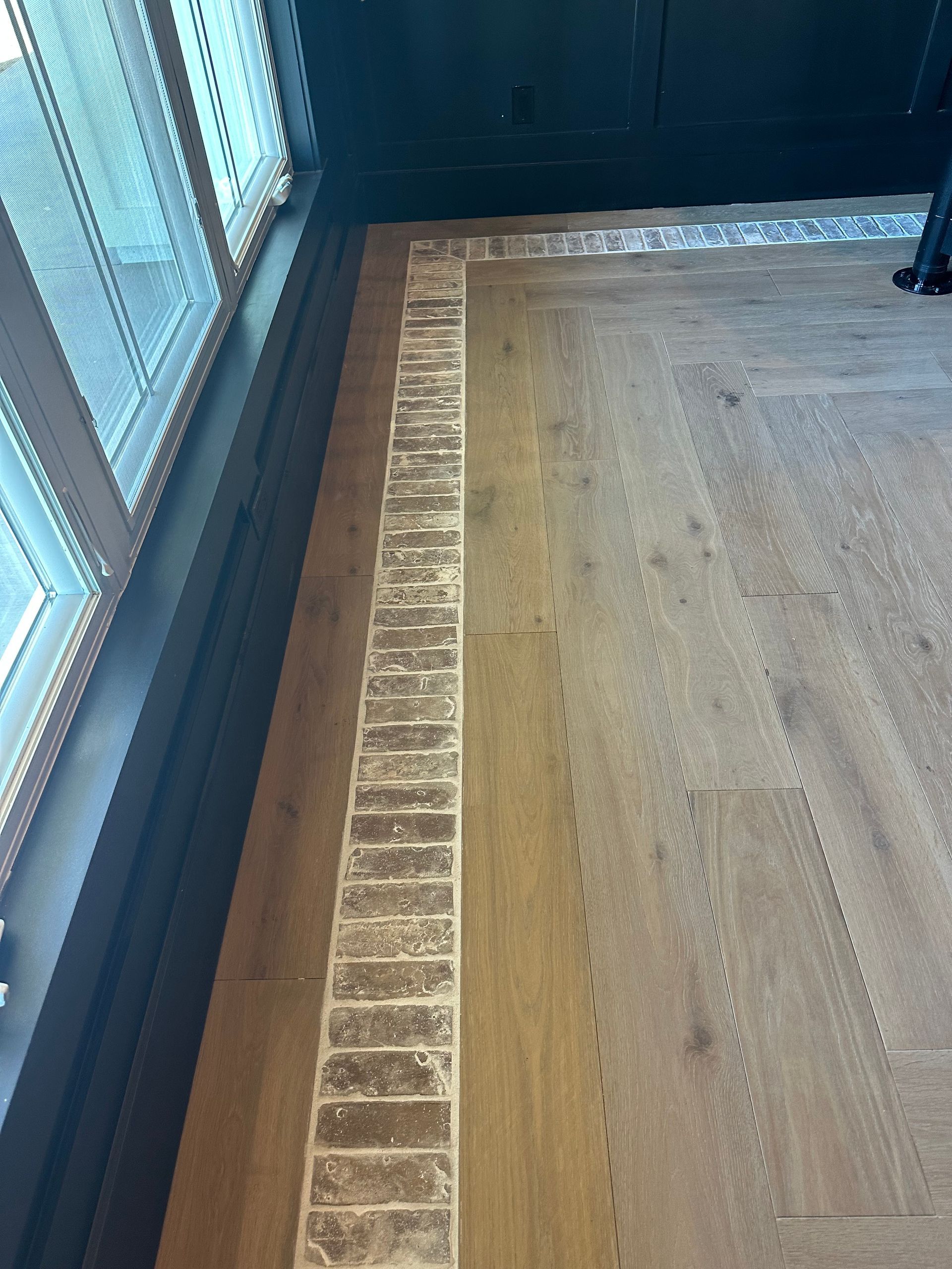 Flooring with light wood planks, a brick border, and a black trim, near a window.