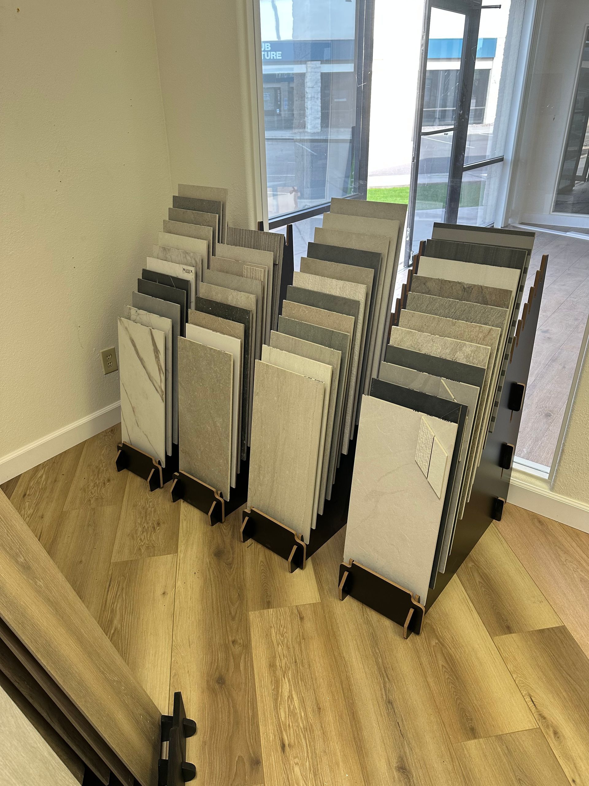 Displays of tile samples on stands in a showroom with a window and wood-look flooring.
