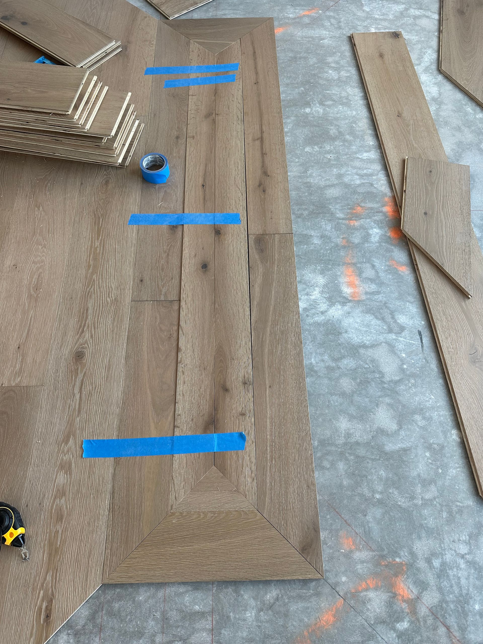 Wooden floor installation in progress; wood planks with blue tape on a gray surface.