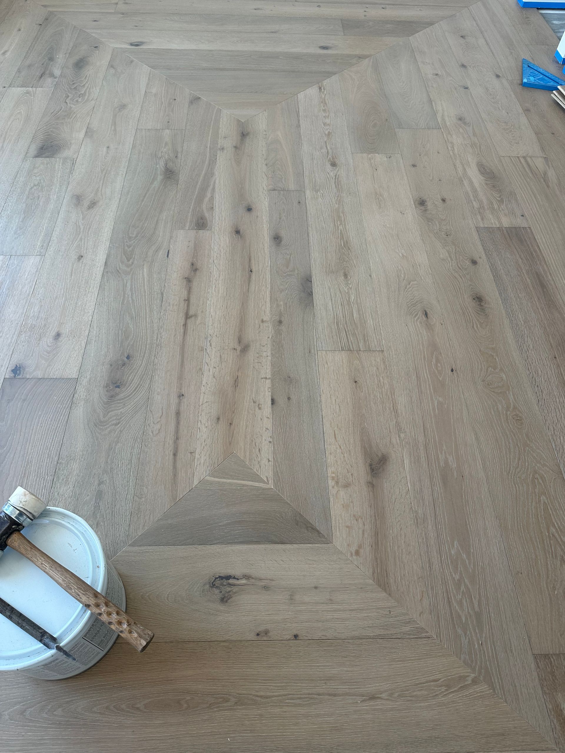 Light-colored wooden floor with a rectangular inlay pattern, a white bucket, and tools nearby.