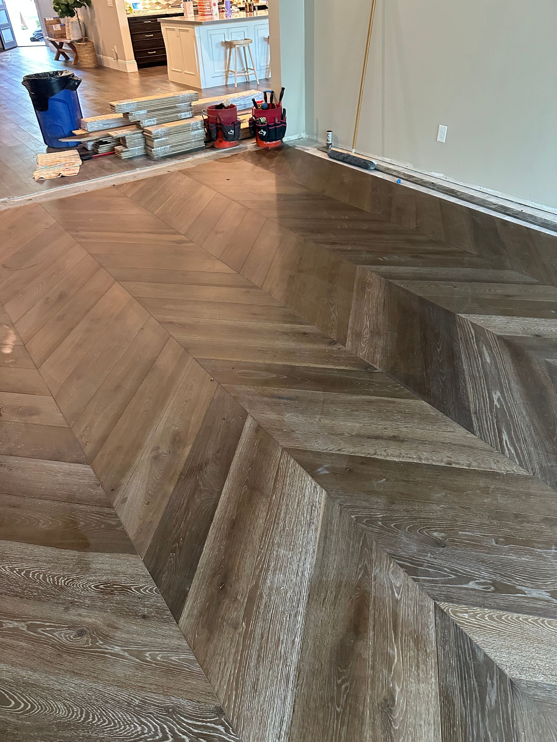 Herringbone-patterned wooden floor in a room, with a kitchen in the background. The flooring is a mix of light and dark brown.