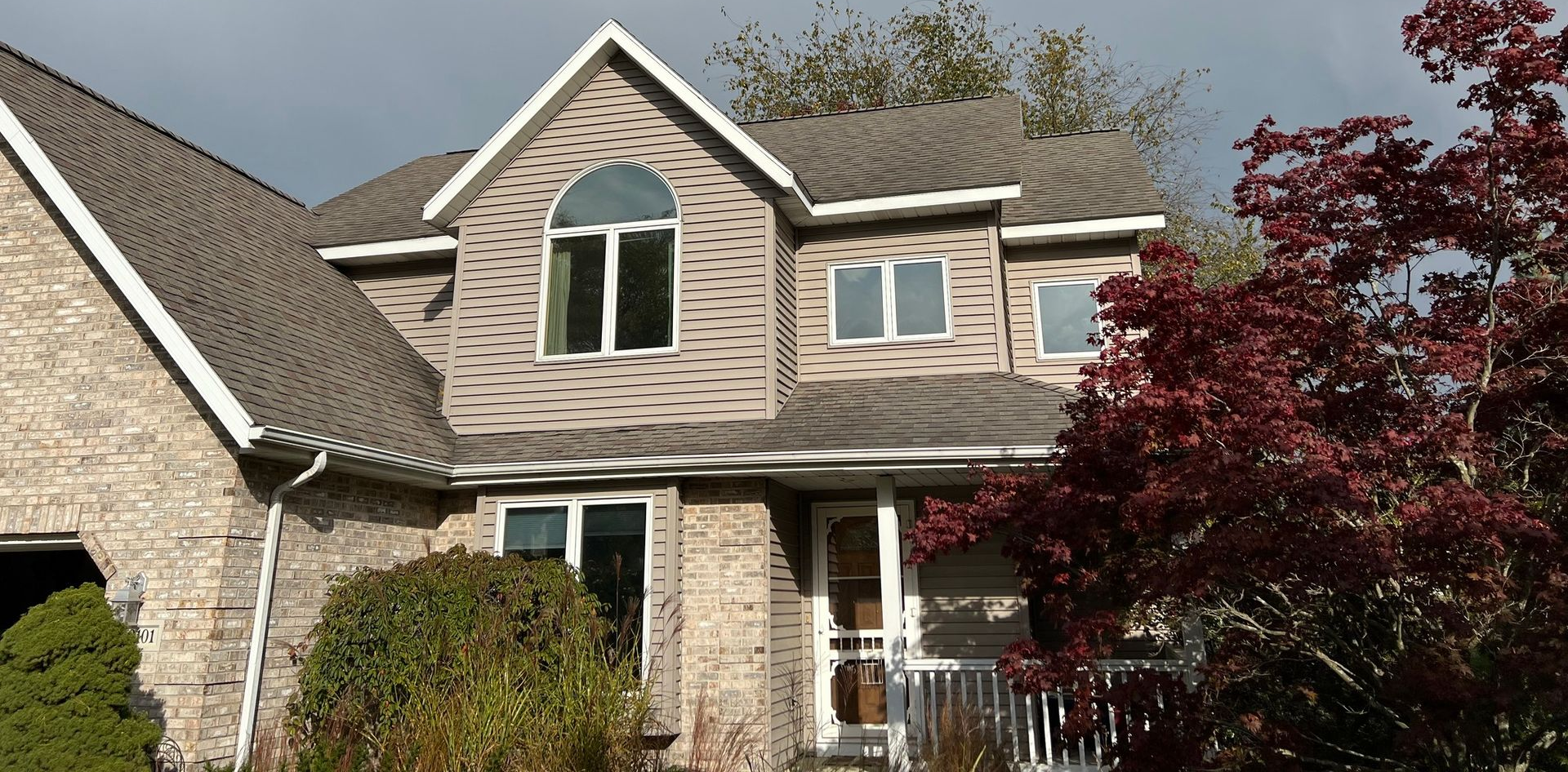 A two-story house with tan siding and a brick facade, trees in the yard.