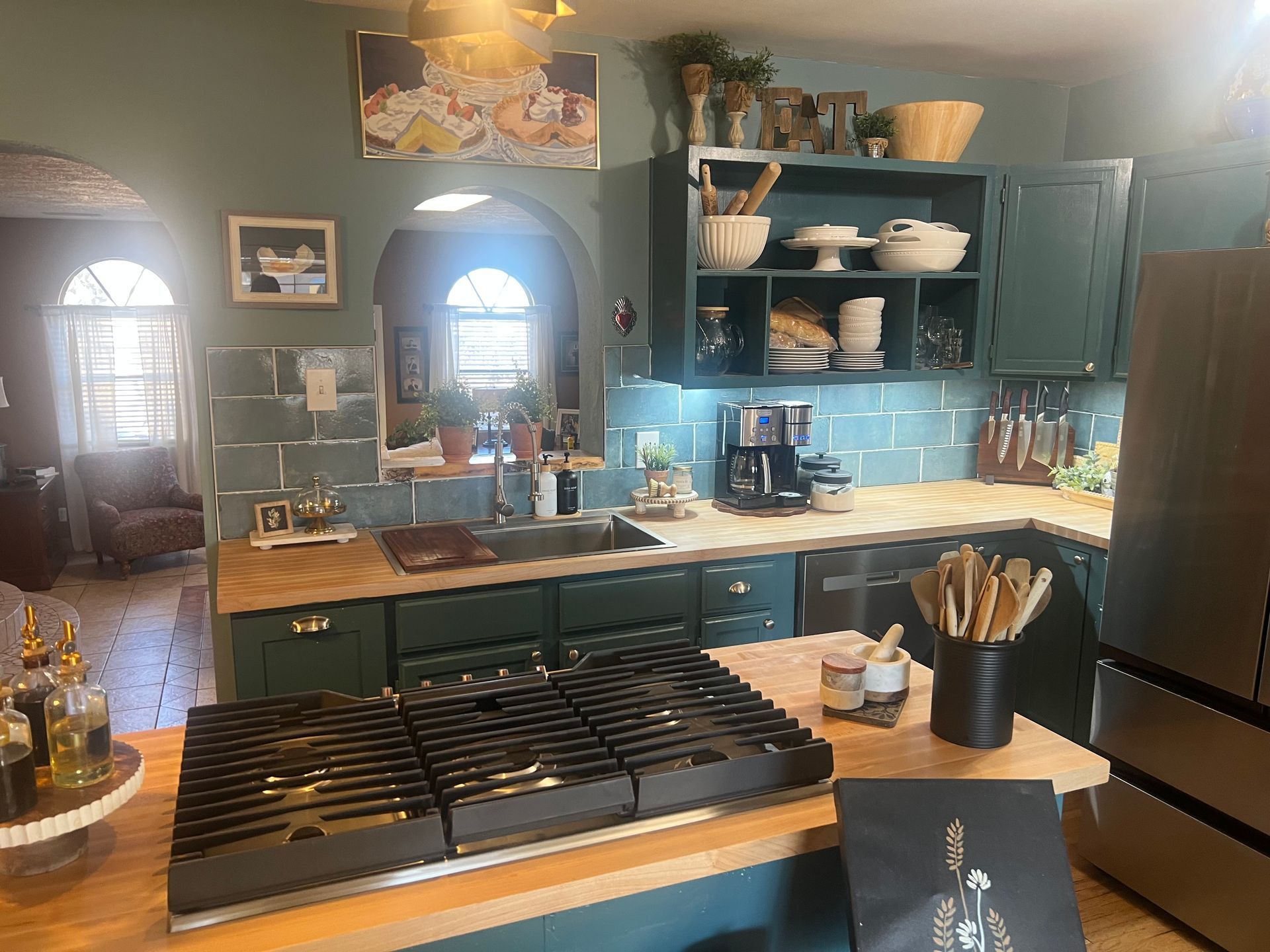 Bright kitchen with dark green cabinets, white island, hanging pots, and a gas stovetop in the foreground.