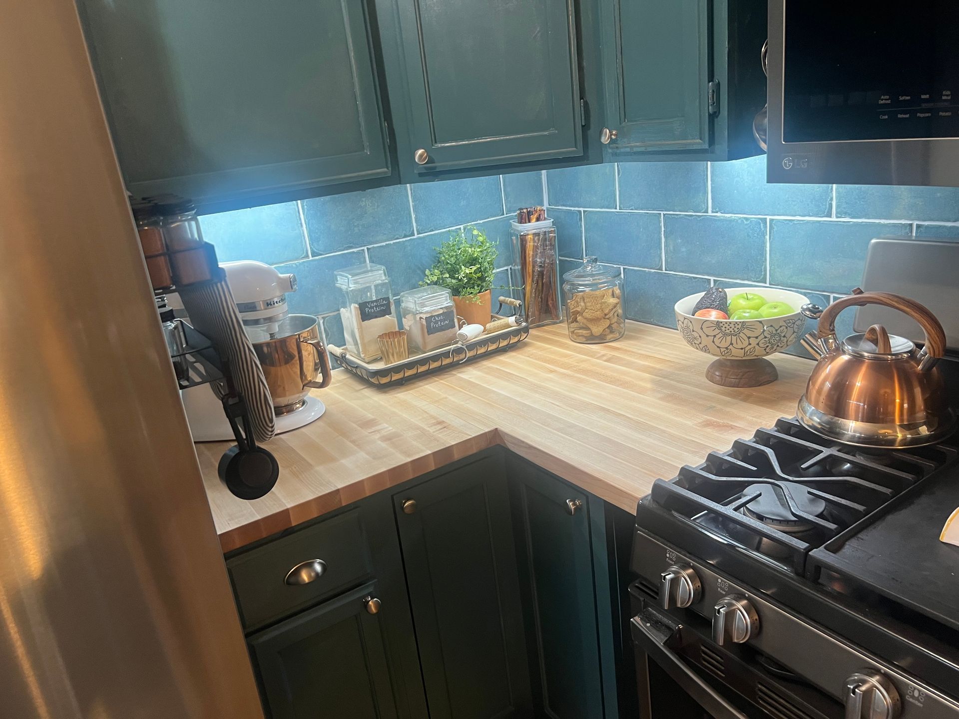 Small kitchen corner with dark green cabinets, striped countertop, stove, kettle, and potted herbs.
