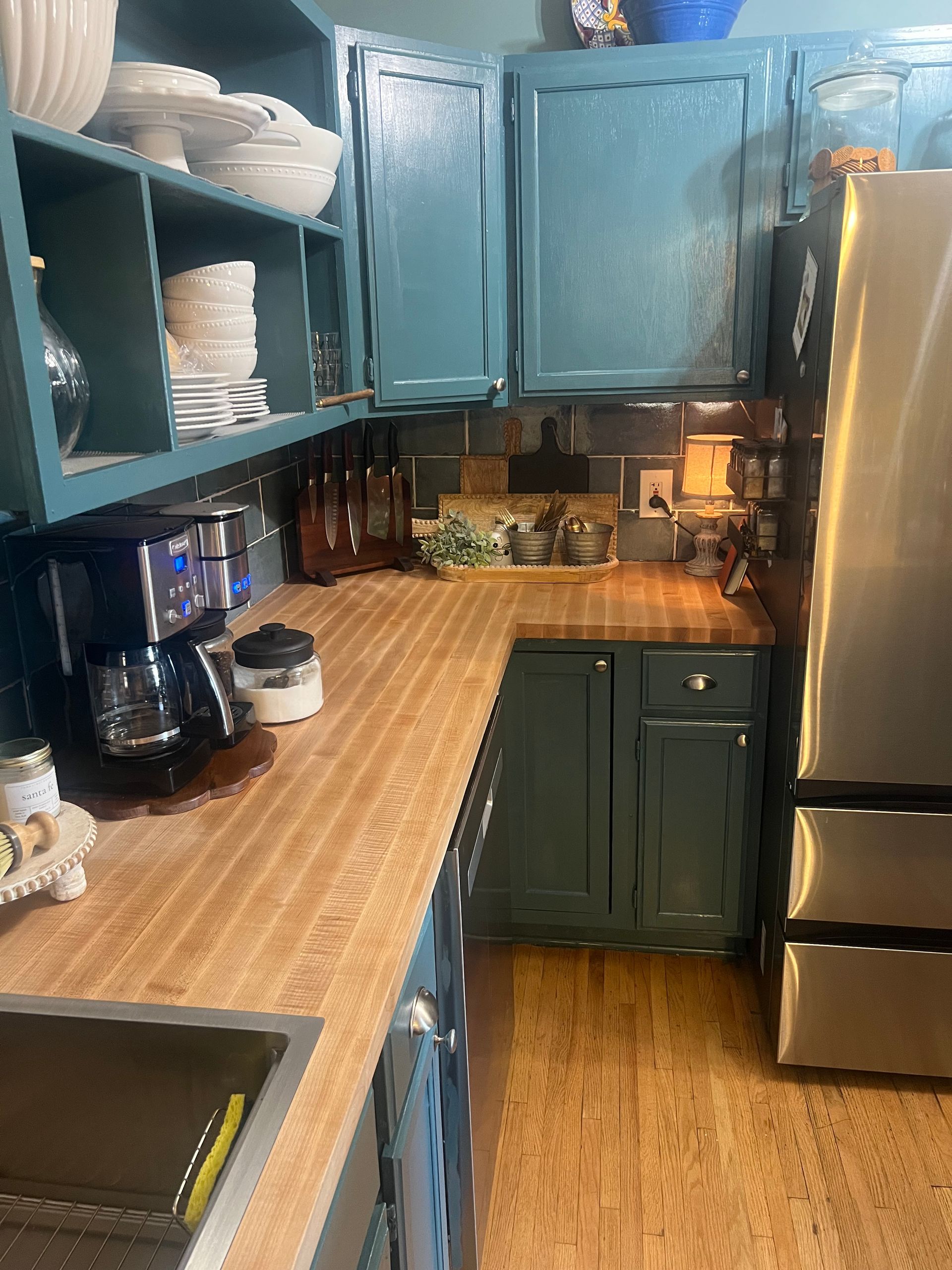 Compact kitchen with teal cabinets, wood countertops, open shelving, and a stainless steel refrigerator.