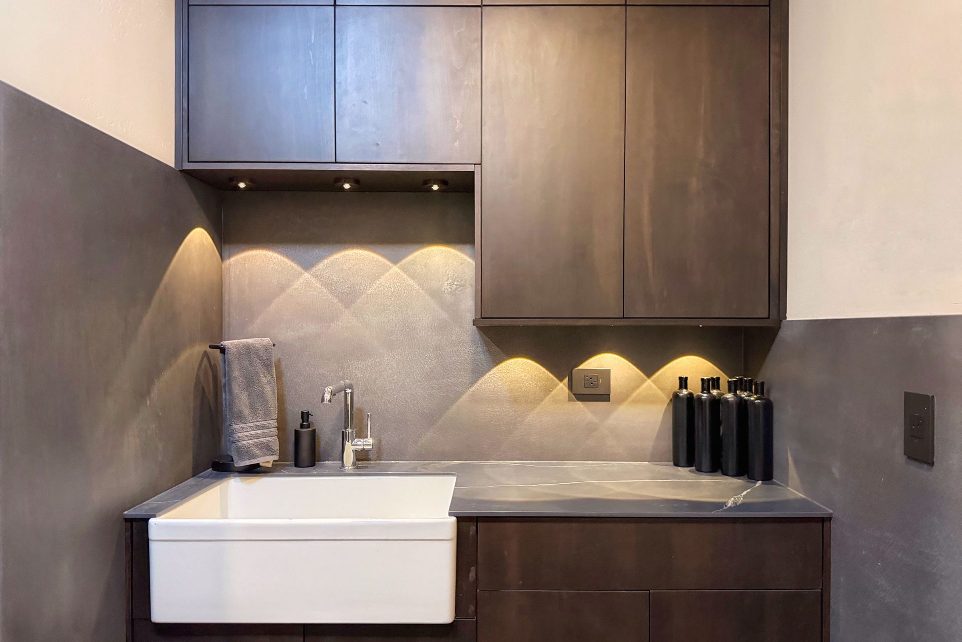 Compact modern kitchen with wood cabinets, gray backsplash, white sink, and under-cabinet lighting