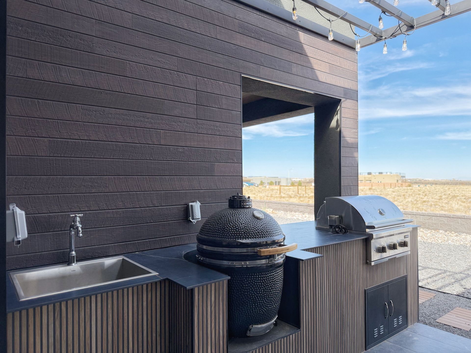 Outdoor patio with sink, grill, and smoker beside a dark wooden wall overlooking a sandy landscape