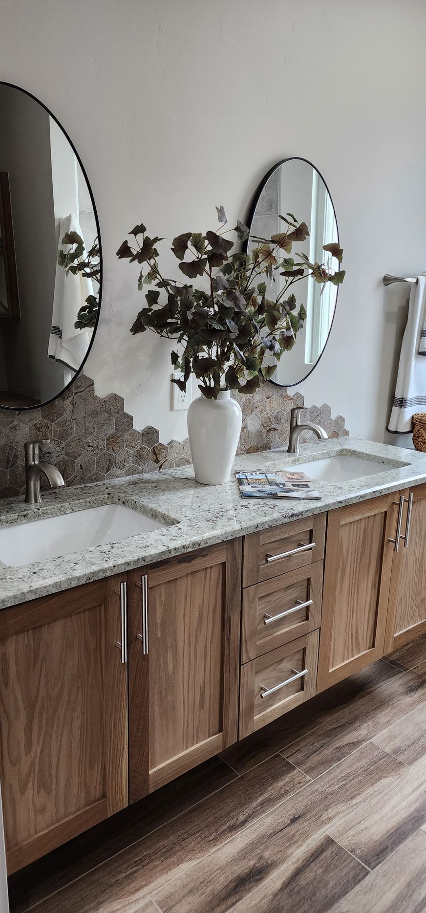 Modern bathroom vanity with wood cabinets, marble countertop, round mirrors, and a white vase with greenery