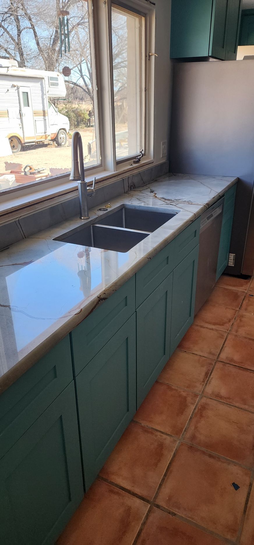 Kitchen with teal cabinets, double sink under a window, and terracotta tile floor