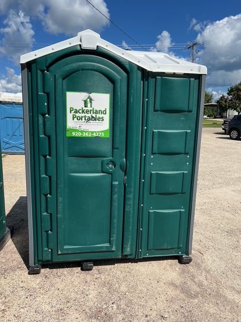 Green porta-potty on a trailer with a cloudy sky background. A road, buildings, and storage tanks are visible.