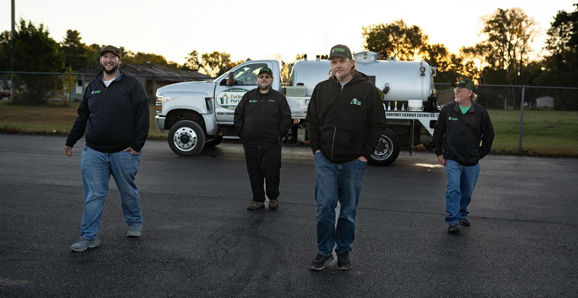 Four men in work attire stand by a truck with a water tank; outdoor setting.
