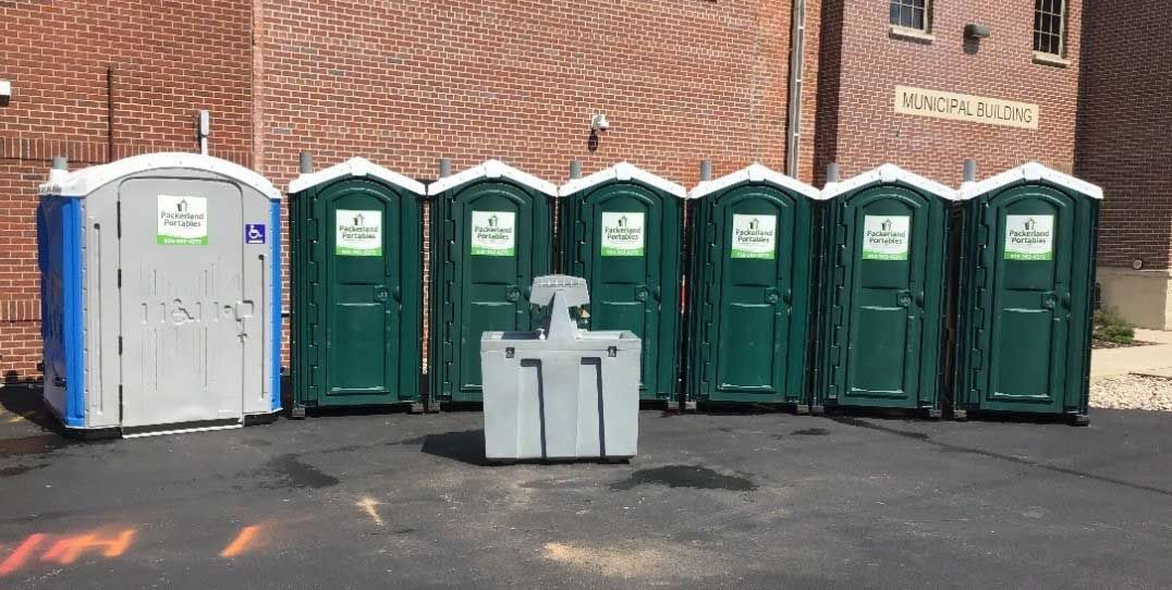 Row of portable toilets of various colors in front of a brick building.