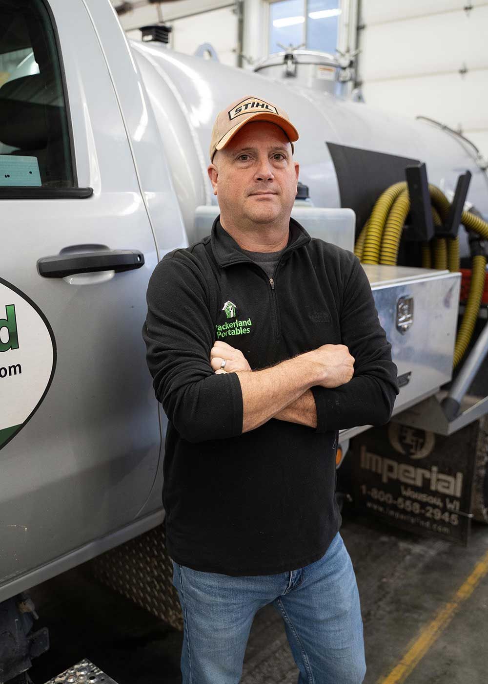 Man in black shirt and cap stands with arms crossed in front of a septic truck.