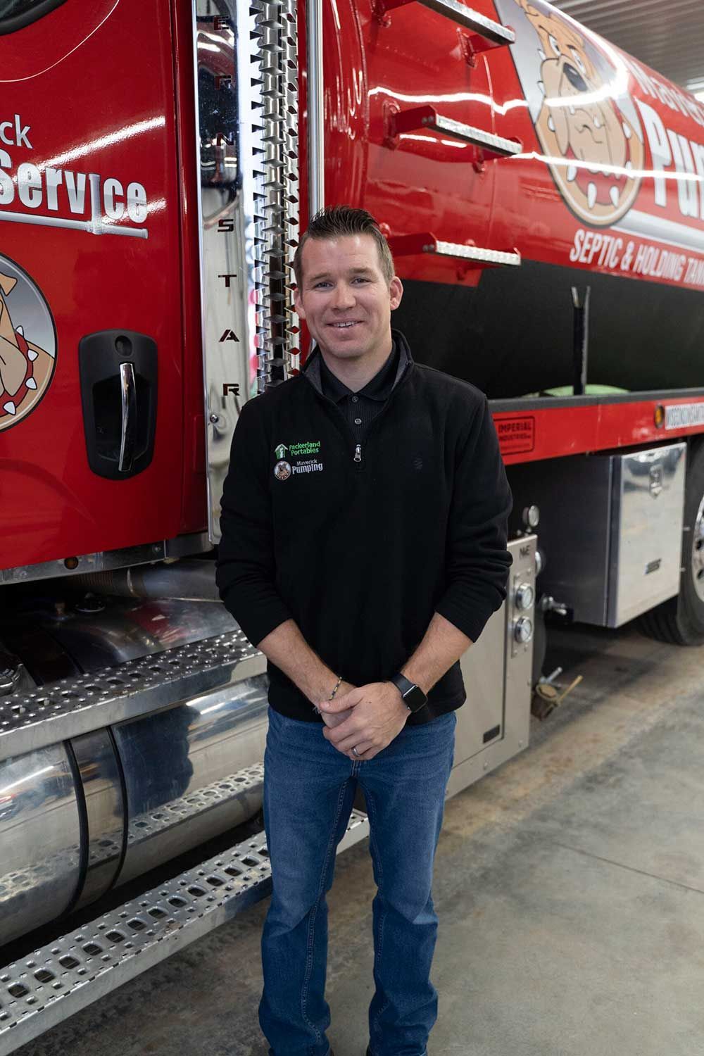 Man in black jacket and jeans stands in front of a red tanker truck.