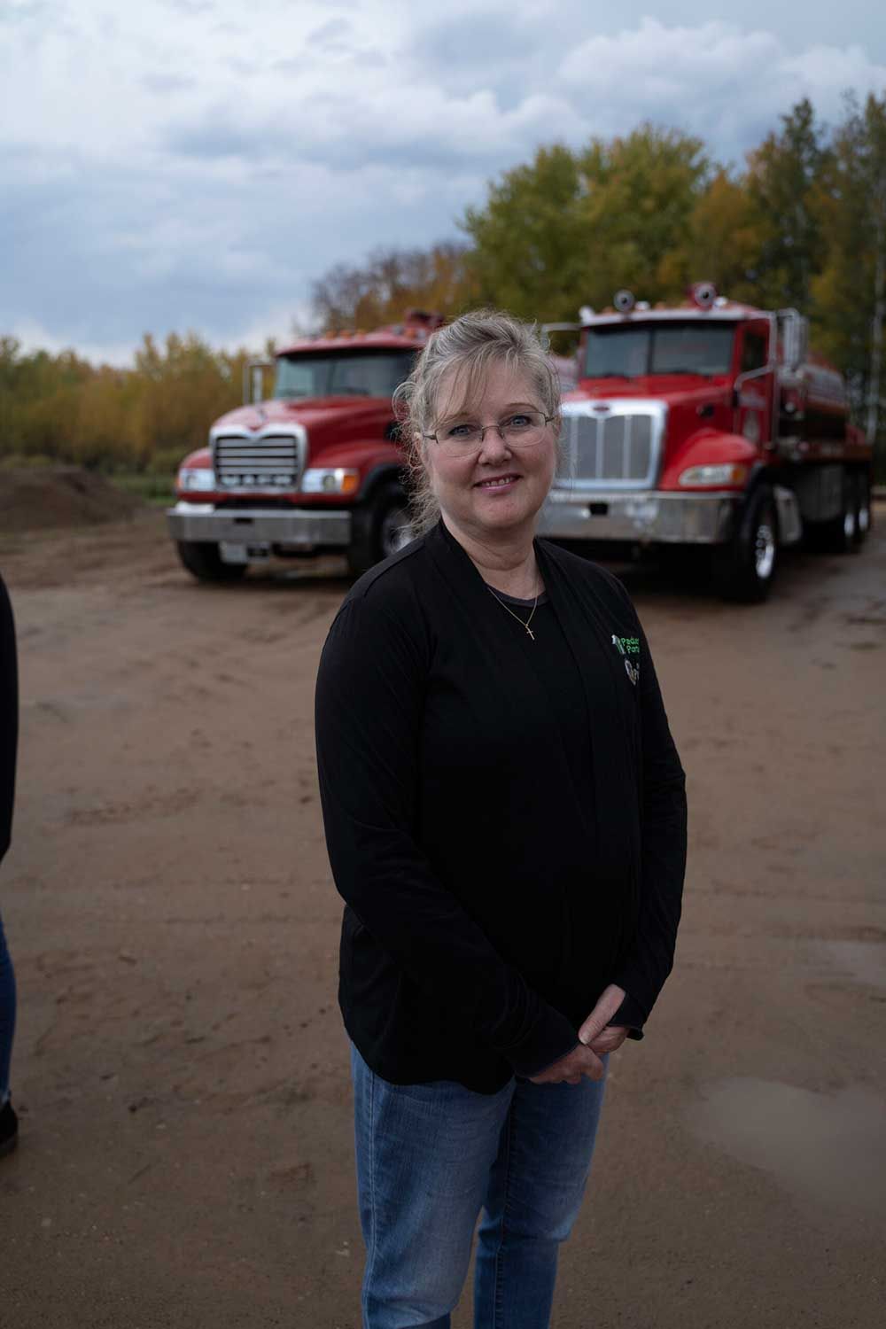 Woman in black shirt and jeans stands in front of two red trucks on a cloudy day.