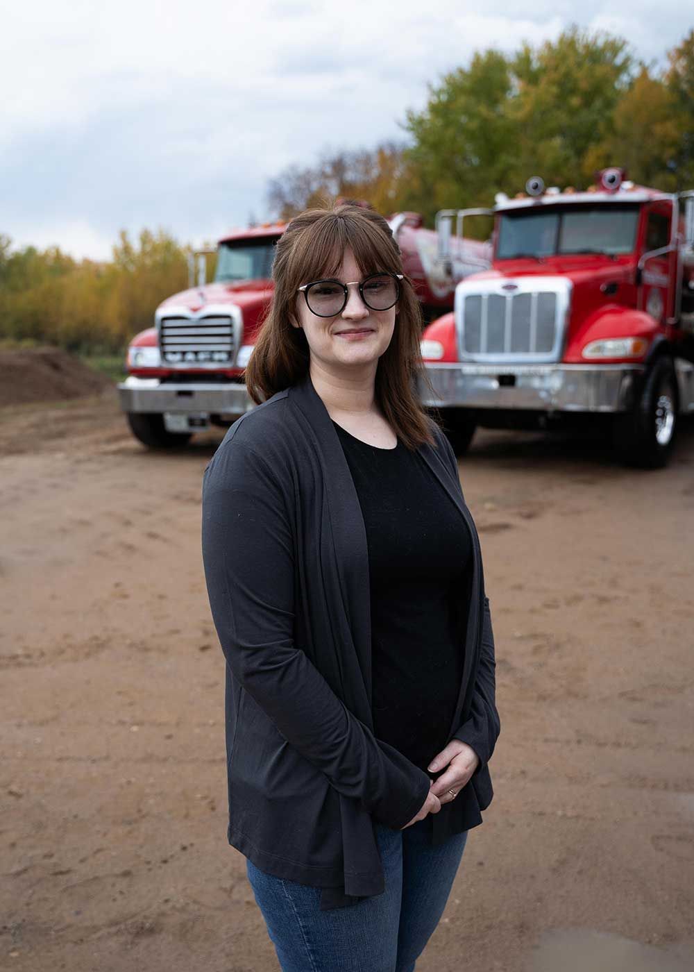 Woman standing in front of two red trucks, wearing a dark cardigan and glasses, outside.
