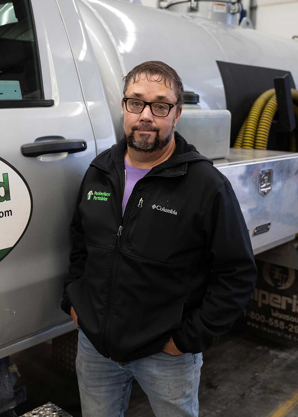 Man with glasses and beard stands by a silver truck wearing a black jacket.