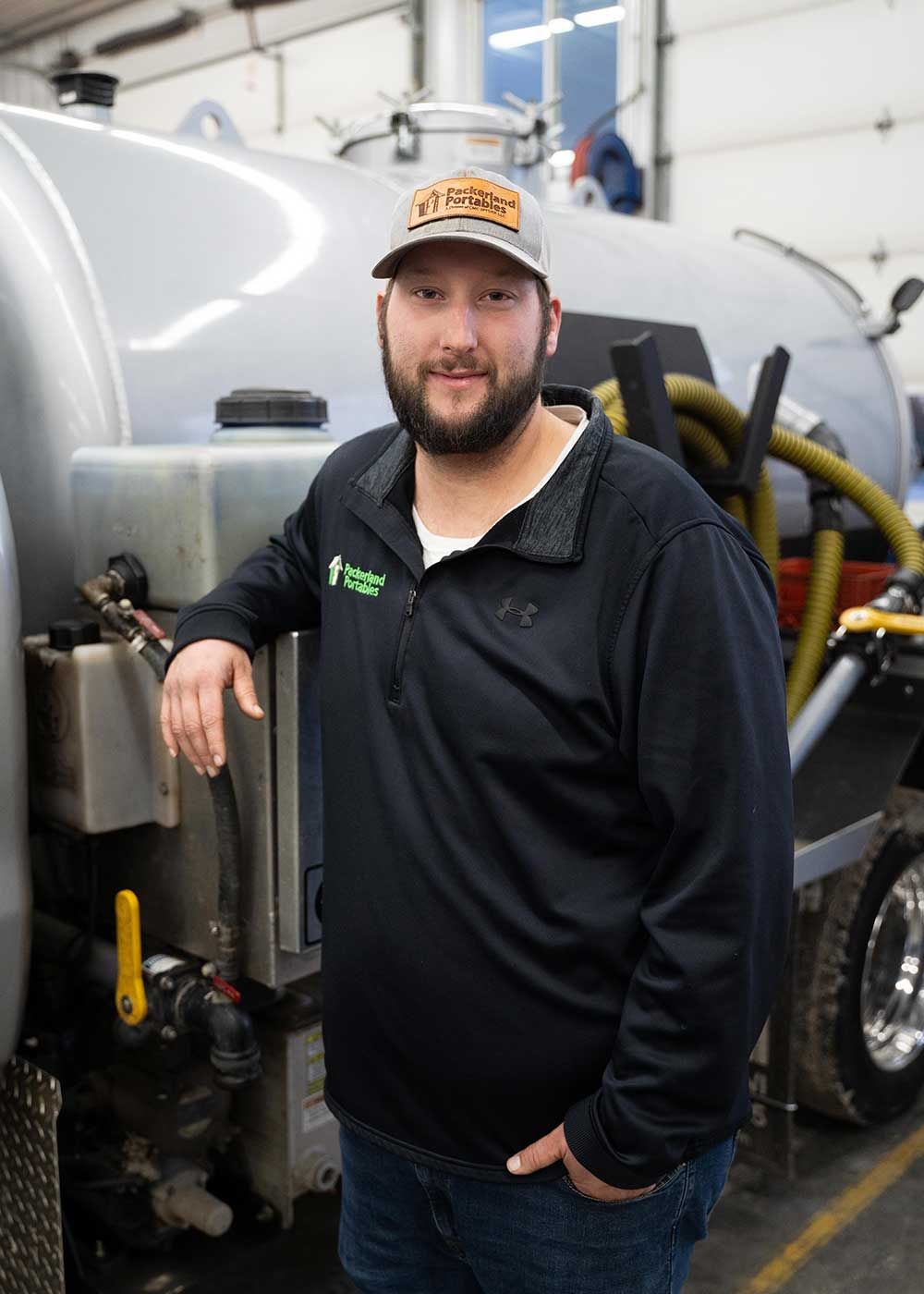 Man in cap, leaning on a large truck, smiles. He wears a black jacket and stands in a garage-like setting.