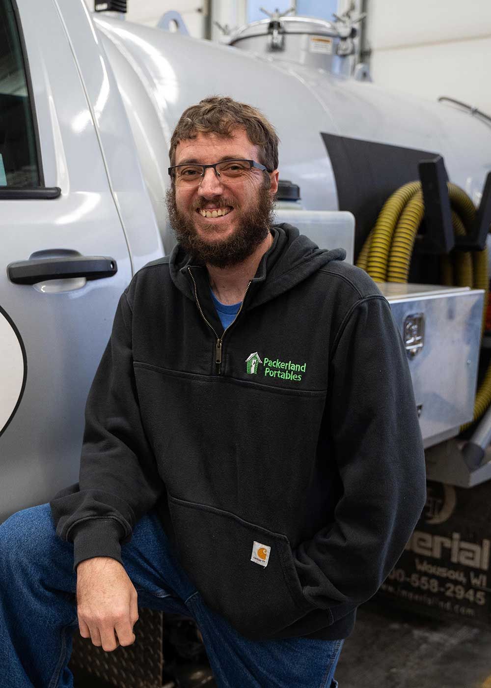 Man in black hoodie with beard smiling, leaning on side of silver truck.