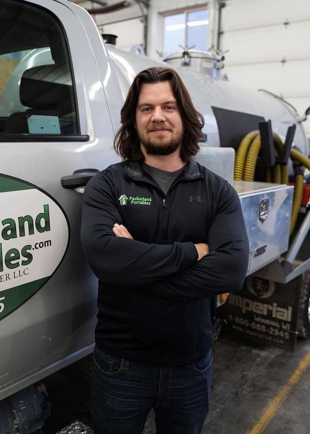 Man with long hair and crossed arms in front of a gray truck. He's wearing a black jacket and jeans.