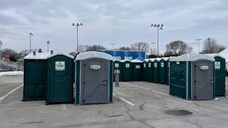 A row of green portable toilets in a gray parking lot, snowy and overcast.