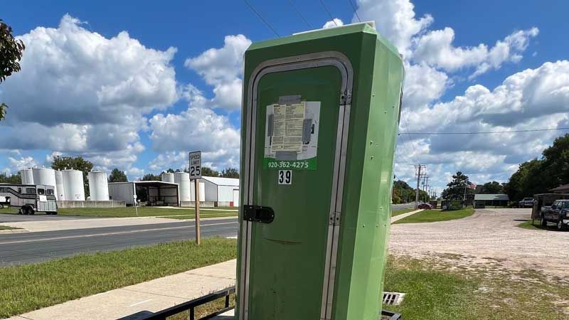 Green porta-potty on a trailer with a cloudy sky background. A road, buildings, and storage tanks are visible.