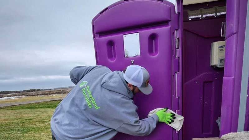 Man in grey jacket cleans the door of a purple portable toilet with a white cloth, outdoors.