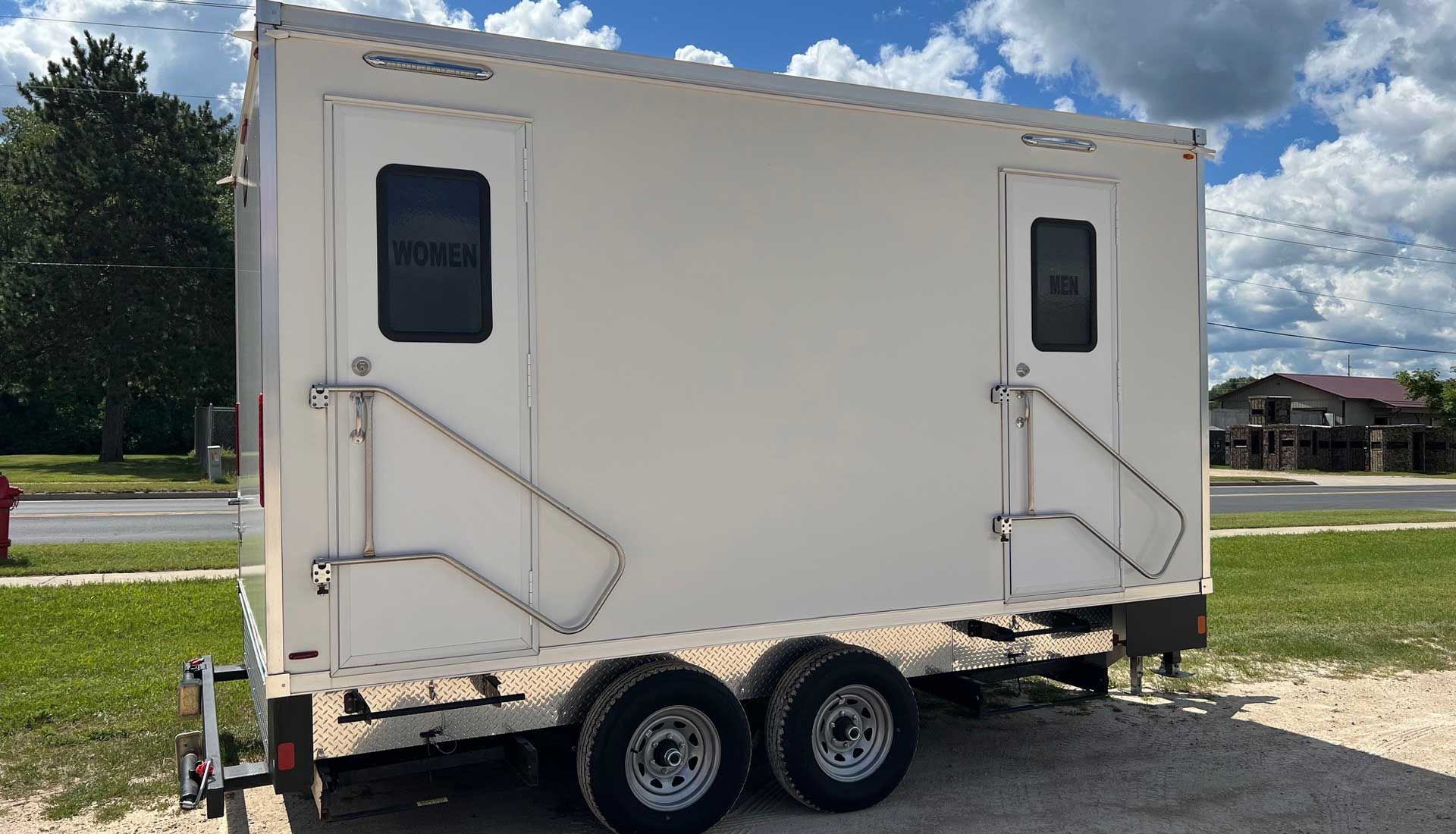Green porta-potty on a trailer with a cloudy sky background. A road, buildings, and storage tanks are visible.