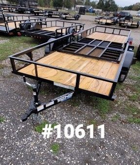 Open utility trailer with wooden deck, metal frame, and surrounding fence, parked outside on gravel.
