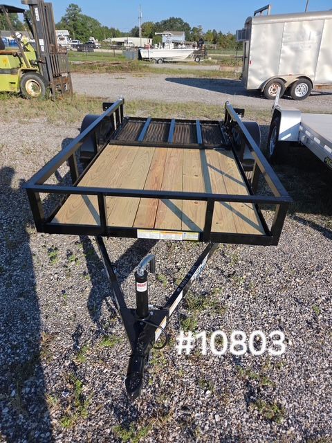 Black metal utility trailer with wooden floor parked on gravel lot, white trailer in background