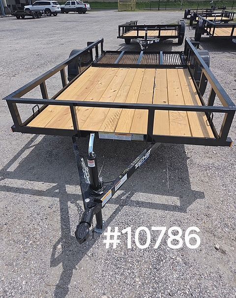 Black utility trailer with wooden deck parked on gravel lot outdoors.