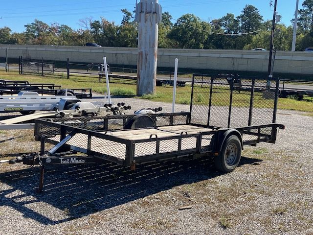 Black utility trailer with mesh sides and a wooden floor, parked on gravel outdoors.