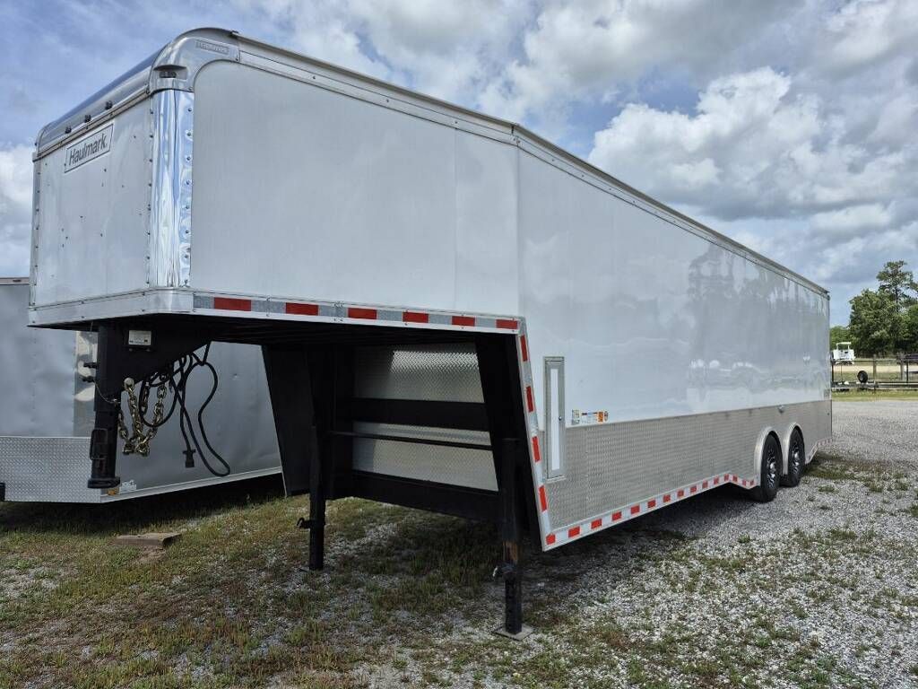 A white, gooseneck-style enclosed cargo trailer parked on a gravel lot under a cloudy sky.