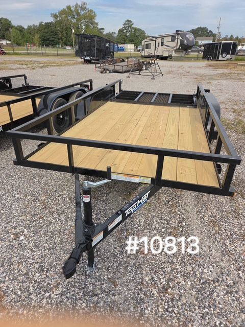 Black flatbed utility trailer with wooden deck parked on gravel lot, other trailers in background.