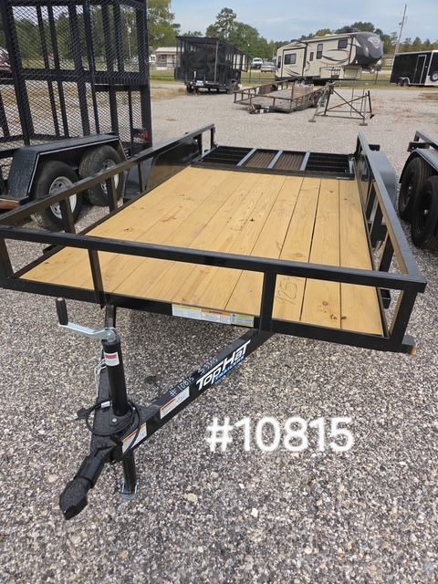 Black utility trailer with wood floor and single jack stand in a gravel lot; other trailers in the background.