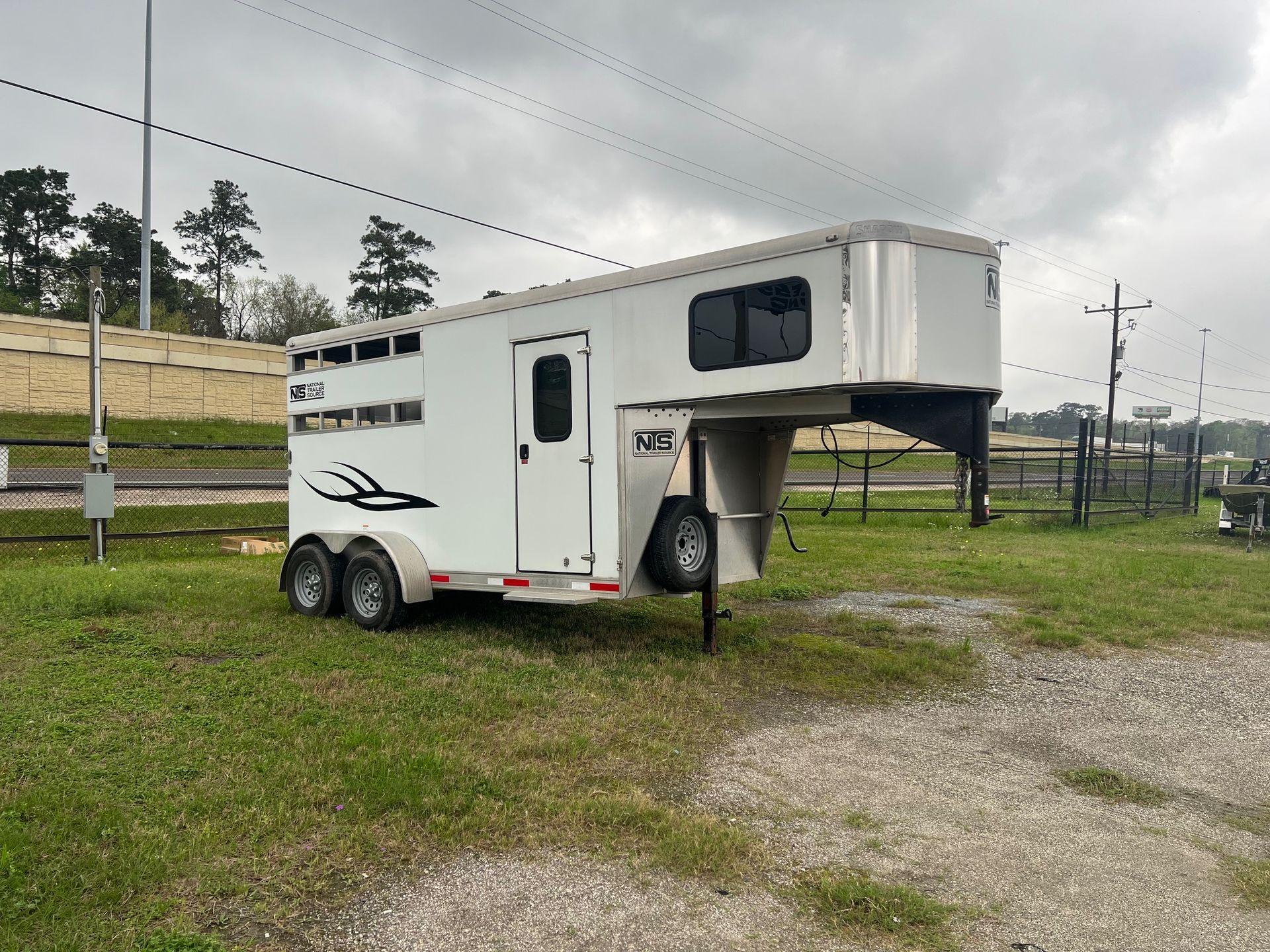 A white gooseneck horse trailer with black accent decals parked on grass in an open, overcast field.