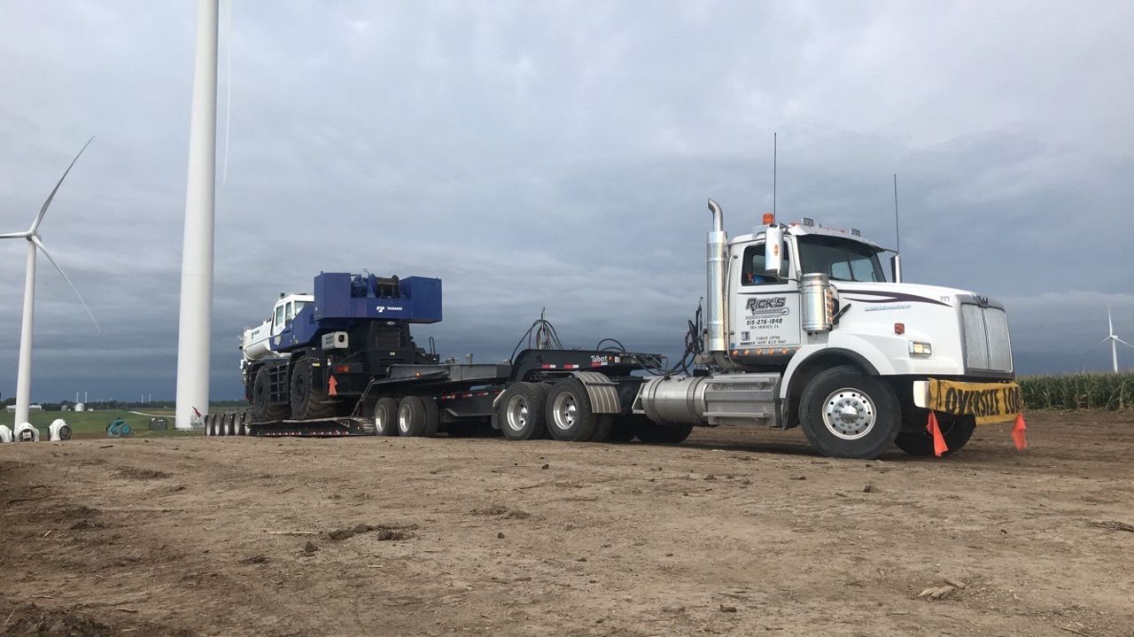 A semi truck is driving down a dirt road next to a wind turbine.