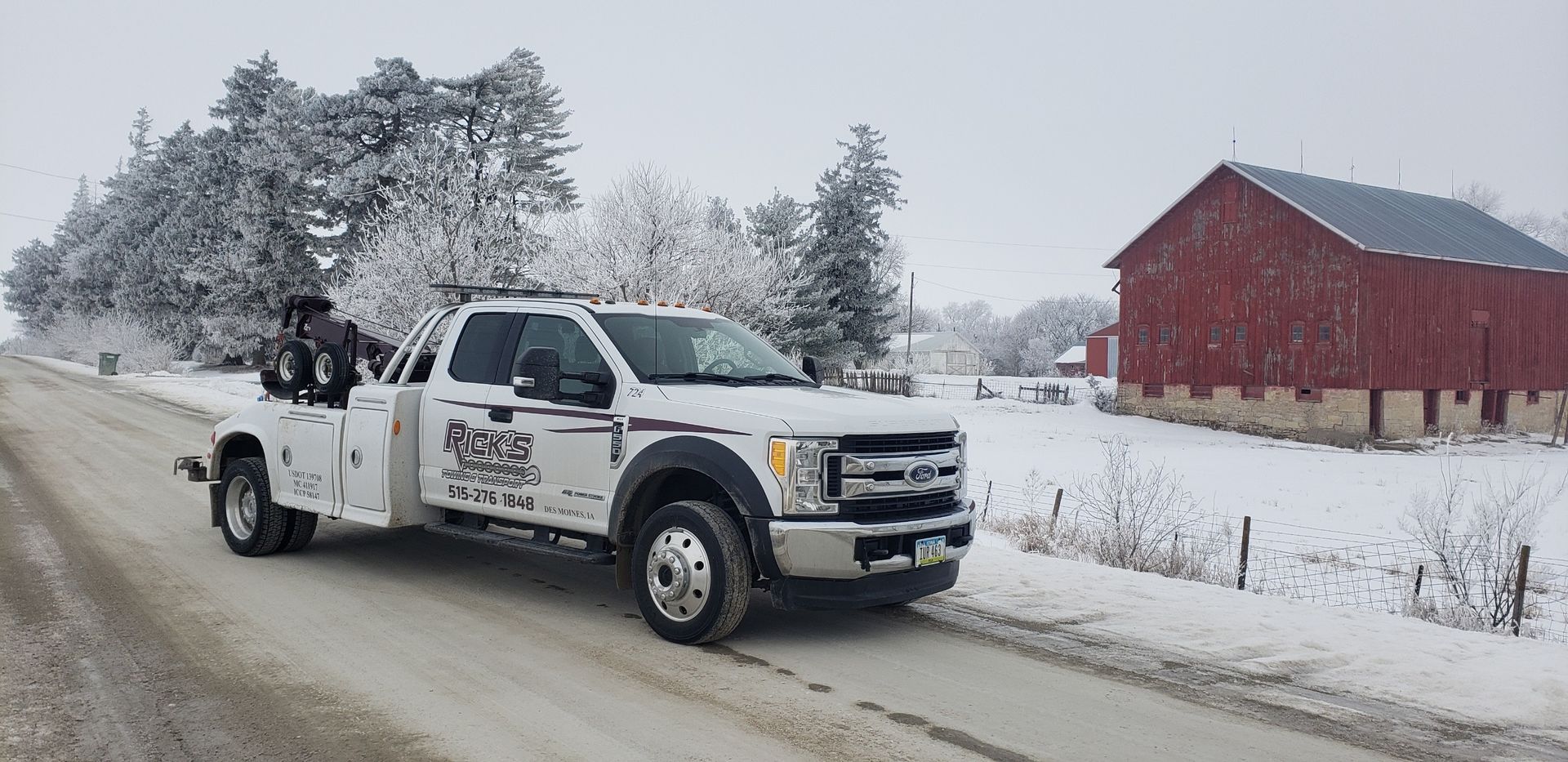 A white tow truck is driving down a snowy road next to a red barn.